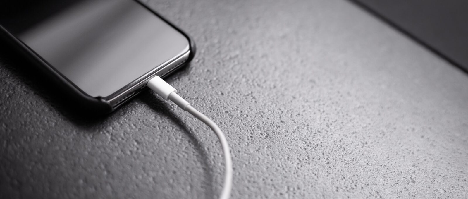 A black iphone resting on a gray slate surface, plugged in.