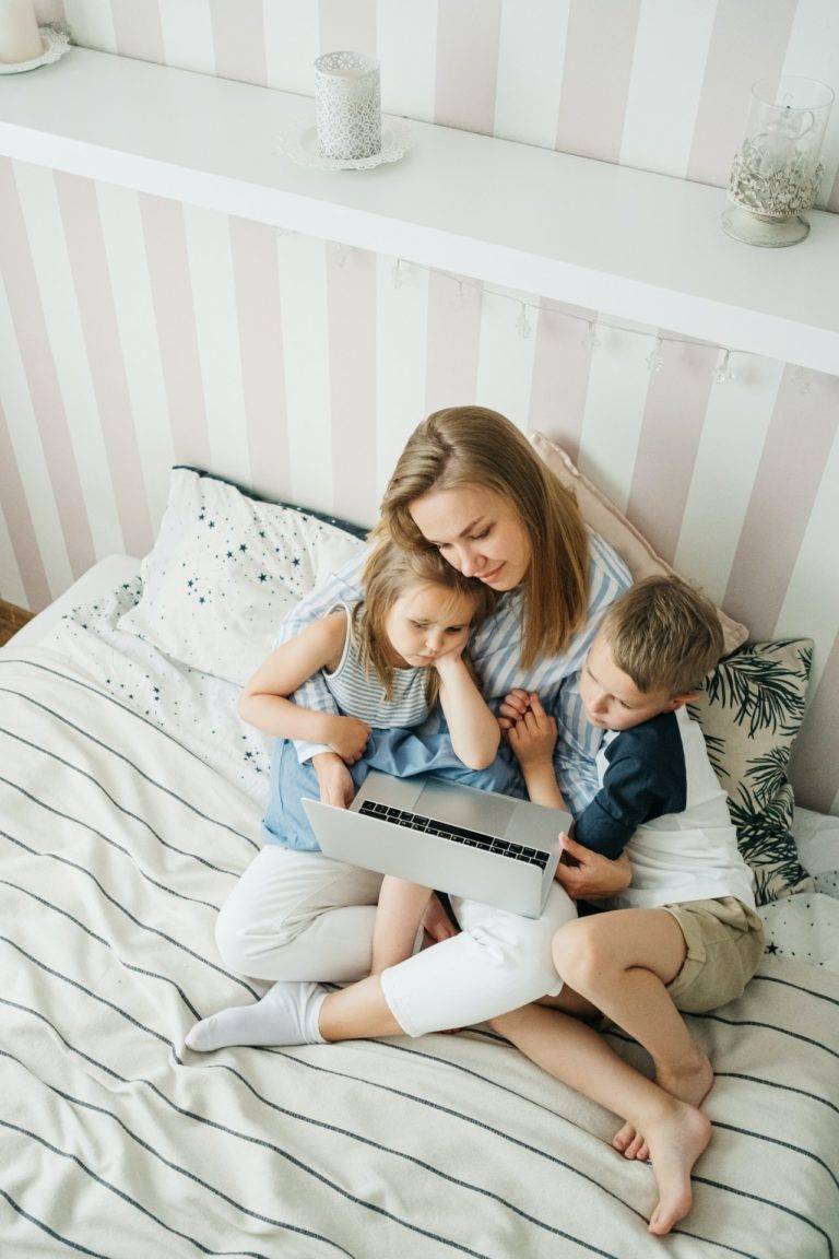 A girl sitting on bed with 2 children using laptop