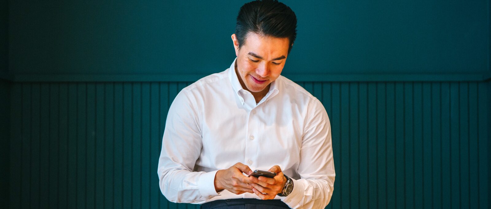 A person in masculine business-casual attire leans against a table while looking down at the phone.